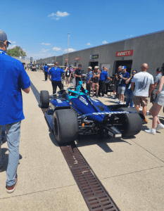 A blue IndyCar is parked in a pit lane surrounded by team members and spectators under a sunny sky near the Andretti garage.